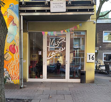 Der mit Gehwegplatten belegte Weg führt mit leichter Steigung vor die Glasfront des Eingangs, in der Mitte die Flügeltür mit der weißen Aufschrift Kulturladen in handschriftlichem Stil, schwer lesbar.