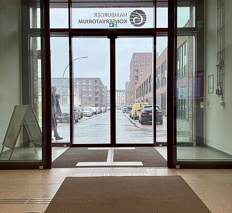 2 goße Glastüren hintereinander. Die innere ist offen, die äußere geschlossen.  Dazwischen auf einer braunen Fläche deutlich abgesetzt helle Leitstreifen. Auf dem Boden vor der inneren Tür ein brauner Teppich, links davon ist ebenfalls ein Leitstreifen zu erkennen. Ganz oben an der äußeren Tür steht Hamburger Konservatorium, hier spiegelbildlich zu lesen. 