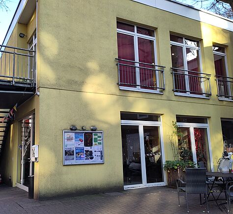 Ein gelbes, 2-stöckiges Haus. An der linken Seitenwand führt eine Feuertreppe auf ein Plateau über der Eingangstür zur "Terassentür" im ersten Stock. Auf der uns zugewandten Wand sind 4 große Fenster mit weißem Rahmen zu sehen. Links an dieser Wand hängt ein Schaukasten, der Inhalt ist nicht zu erkennen. Auf dem kleinen Platz vor dem Haus stehen ein Stuhl und ein Tisch.