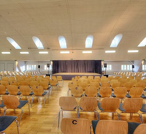Bestuhlter Saal, Blickrichtung Bühne mit zugezogenem Vorhang. Hohe gewölbte Decke mit hufeisenförmigen Fenstern über der Bühne