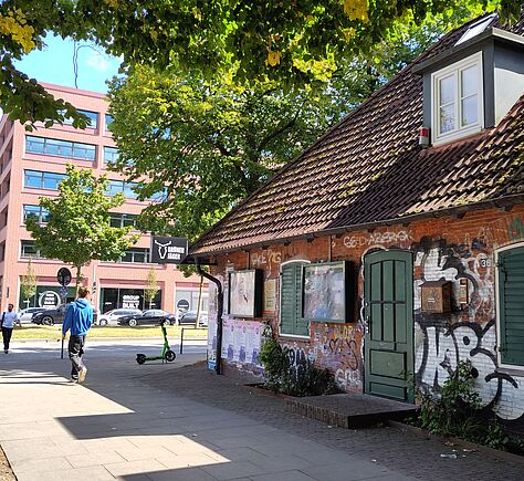 Ein kleines Backsteingebäude mit schrägem Dach und einer Gaube steht rechts von einem breiten Gehweg. Die Fassade ist mit Graffiti bedeckt, zwei Schaukästen hängen an der Wand. Eine grüne Tür und zwei mit grünen Läden geschlossene Fenster sind sichtbar. Vor der Tür ist eine niedrige Stufe. Über dem Haus spenden große Bäume Schatten. Im Hintergrund ist ein modernes Gebäude mit roter Fassaden und viele Fenstern. Auf dem Gehweg gehen Menschen, ein E-Scooter steht am Straßenrand.
