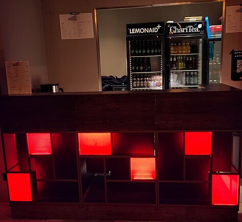 Im Vordergrund eine dunkle Theke, darunter ein Regal mit mehreren Fächern; einige Flächen leuchten in warmem Rot. Dahinter stehen zwei Getränkekühlschränke. Auf der Theke steht eine Getränkekarte.