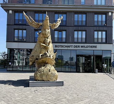 Auf einer gepflasterten freien Fläche steht eine große, goldfarbene Skulptur mit tierischen Formen auf einem Sockel mit spiralförmigen Mustern. Im Hintergrund befindet sich ein modernes Gebäude mit dunkler Fassade und großen Fenstern. Über dem Eingang steht in schwarzen Buchstaben ‚Botschaft der Wildtiere‘.