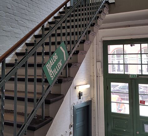 Von der Eingangstür führt ein Treppenhaus zu einer weiteren grünen Tür. Die Treppe hat in Richtung aufwärts rechts einen Handlauf. Die Wände sind weiß. Seitlich am Geländer ein Schild: DIALOG IM STILLEN.