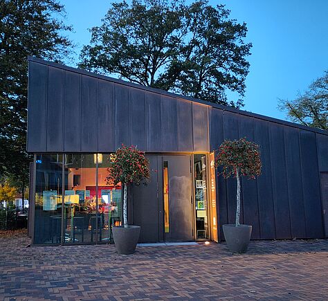 Modernes Gebäude mit dunkler, schräger Fassade. Am Eingang stehen zwei große Pflanzkübel mit kleinen Bäumen. Links neben der Tür ist eine Glasfront, durch die den beleuchteten Innenraum sieht. Innen Lichtquellen und Menschen. Vor dem Gebäude befindet sich ein gepflasterter Platz, im Hintergrund sind Bäume und blauer Himmel bei Dämmerung. 