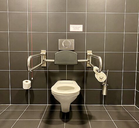 Das Rollstuhl-WC ist dunkelgrau gefliest, das WC-Becken weiß. Links und rechts viel Platz neben dem WC, zwei Stützklappgriffe. Das WC ghat eine gepolsterte Rückenlehne.