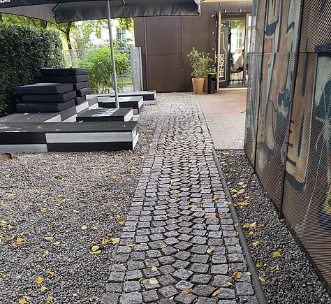 Ein gepflasterter Weg führt durch einen Außenbereich, rechts und links vom Weg grauer Kies. Links vor einer Hecke Podeste mit schwarzen Polstern unter einem Sonnenschirm. Rechts vom Weg ist auf der Metallverkleidung des Gebäudes farbige Graffiti.