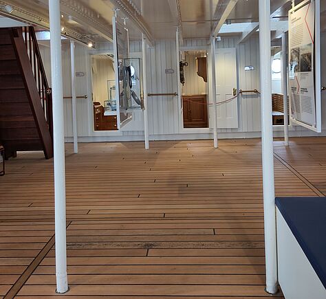 Holzvertäfelter Innenraum mit weißen Metallstreben, die den Raum in regelmäßigen Abständen gliedern. Zwischen den Streben hängen Ausstellungstafeln mit Text und Bildern. Links führt eine dunkle Holztreppe nach oben. Der Boden besteht aus hellem Holz. Im Hintergrund sind drei geöffnete Türen, die in kleine Räume führen. Vorne rechts ist eine blauweiße Bank. Licht fällt von oben. Die Atmosphäre wirkt sachlich und aufgeräumt.