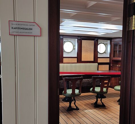 Das Foto zeigt eine geöffnete Tür mit dunklem Holzrahmen, hinter der sich ein Raum mit hellem Holzboden und weißer Decke befindet. Die Wände sind aus dunklem Holz mit hellen Fächern. Im Zentrum stehen ein länglicher Tisch mit roter Kante und mehrere Stühle mit grün gepolsterten Sitzen. An der Wand befindet sich eine gepolsterte Sitzbank, darüber sind zwei Bullaugen. Links neben der Tür hängt ein Schild mit der Aufschrift 'KAPITÄNSSALON'.