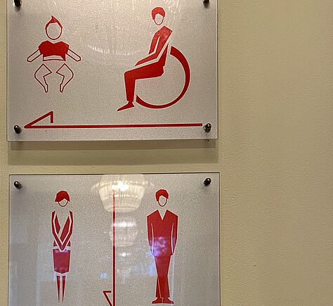 Zwei weiße Schilder mit roten Piktogrammen. Das eine für Rollstuhl-WC mit Wickeltisch. Ein Pfeil nach links zeigt die Richtung an. Das andere Schild für das Männer und Frauen WC. Der Pfeil zeigt nach unten.