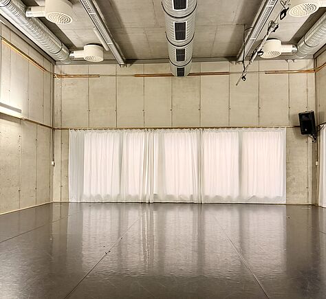 Ein leerer, heller Saal mit glattem, dunklem Boden und hohen, cremefarbenen Wänden. An der Stirnseite sind bodentiefe Fenster mit weißen, halbtransparenten Vorhängen. Die Decke ist mit sichtbaren Lüftungsrohren und runden Lüftungsauslässen versehen. In der rechten Ecke hängt ein Lautsprecher, an der Decke verlaufen Schienen mit Scheinwerfern. Der Raum wirkt modern und funktional.