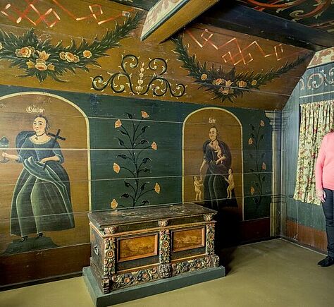 Dunkelgrün und braun verzierte Wände, zwei Gemälde von Frauen an einer Wand, davor eine große Truhe. 2 Besucherinnen am rechten Rand