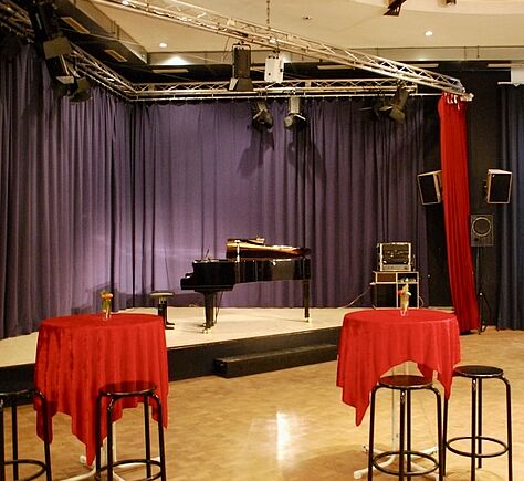 Die Bühne, im Hintergrund ein geschlossener schwarzer Vorhang. Auf der Bühne steht ein schwarzer Flügel, vor der Bühne, zwei hohe Tische mit roten Tischdecken und Barhocker an. Über die Bühne Scheinwerfer.