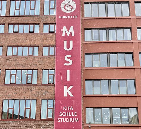 Eine rotgeklinkerte Fassade eines 5-stöckigen Hauses mit Reiehn von Fenstern. In der Mitte hängt ein rotes Banner mit folgendem Inhalt: Ganz oben das Logo in weiß (ein Baßschlüssel), darunter horizontal geschrieben HHKON.DE, darunter vertikal in sehr großen Buschstaben MUSIK. Darunter in 3 Zeilen horizontal die Wörter KITA, SCHULE, STUDIUM