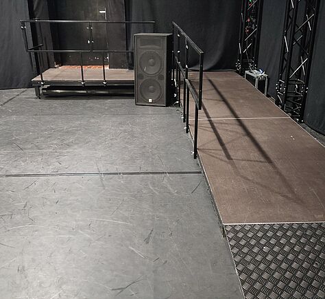 Rampe auf dem dunklen Boden. Die Rampe führt nach oben auf eine kleine erhöhte Plattform mit einem schwarzen Geländer. Links neben der Rampe steht ein großer Lautsprecher.