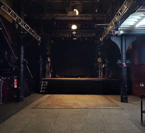 Circa 3 Meter vor der Bühne links und rechts alte Stahlsäulen. Geriffelter Betonboden, nur direkt vor der Bühne liegen Sperrholzplatten. Der Raum ist dunkel, Scheinwerfer an der Decke