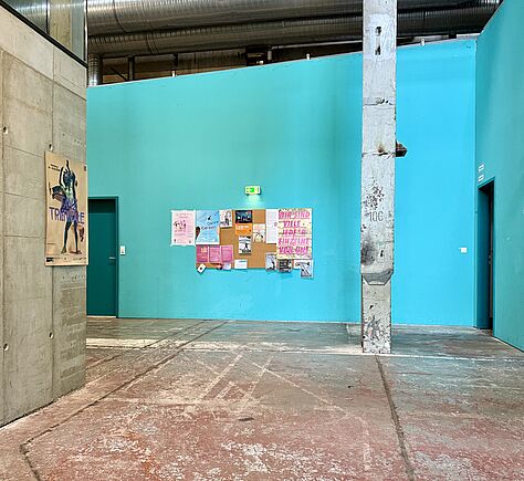 Ein hoher, industriell wirkender Raum mit türkisfarbener Wand. An der Wand hängen mehrere bunte Plakate und Zettel. Nach links geht der Gang weiter. Der Boden ist abgenutzt und rötlich grau. Rechts steht eine viereckige Betonsäule, links an der grauen Wand ist ein Plakat mit einer tanzenden Figur. Links und rechts befinden sich zwei Türen in dunklerem Türkis. Unter der Decke verlaufen silberne Lüftungsrohre.