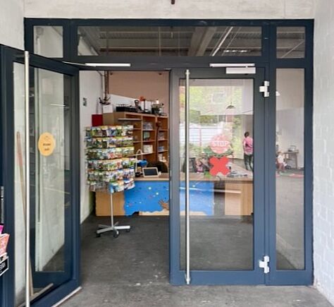 Die Doppeleingangstür ist aus Glas mit dunkelblauem Rahmen. Die Griffe sind senkrecht. Auf dem Foto ist die linke Tür geöffnet, dahinter ist der niedrige Empfangstresen und ein Ständer mit kleinen Büchern.