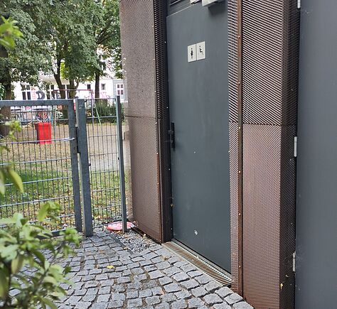 Eine graue Tür mit zwei Piktogrammen, die auf eine barrierefreie Toilette und die für Frauen hinweisen, befindet sich an einem modernen Gebäude mit Metallverkleidung. Der Zugang ist über einen gepflasterten Weg möglich, der direkt zur Tür führt. Links im Vordergrund sind einige grüne Blätter, hinter einem Metallzaun sind Bäume, ein öffentlicher Platz und Häuser erkennbar.