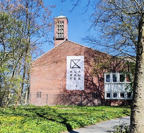 Das Backsteingebäude trägt ein großes weißes „Klabauter“-Schild an der Fassade. Davor führen Wege durch Grünflächen und hohe Bäume rund um das Haus.