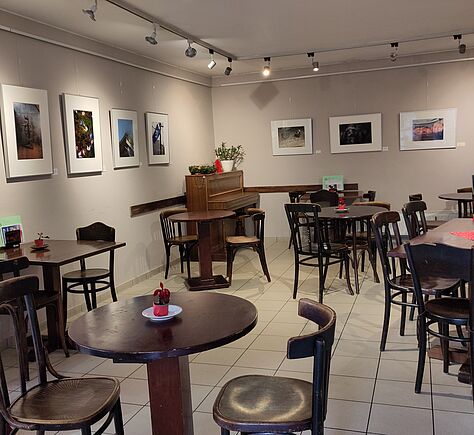 Ein weiß gestrichenener Raum mit Cafétischen und Stühlen. An den Wänden hängen auf einer Höhe viele Bilderrahmen, am Rand der Decke hängen Lampenleisten, die die Bilder beleuchten.