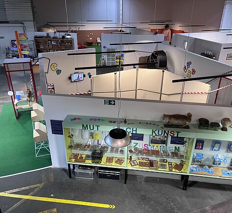 Blick von der Galerie auf die Ausstellungsfläche in einer Halle mit mehreren Trennwänden. Im Vordergrund steht eine Vitrine mit verschiedenen von Kindern gebastelten Objekten. Es gibt eine Vorrichtung für Flaschenpost. Im Hintergrund lehnt eine große Puppe an der Wand.
