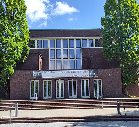 Großes Backsteingebäude mit hohen Fenstern und einer  breiten Treppe vor dem Eingang, mittig 2 Handläufe aus Metall.  links und rechts stehen 2 hohe Bäume.