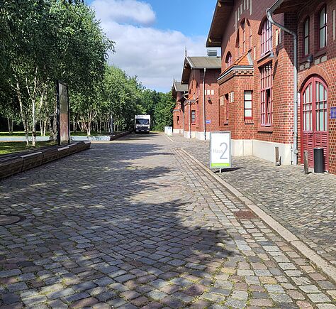 An einer gepflasterten Straße und einem gepflasterten Fußweg stehen mehrere Backsteingebäude mit Holzgiebeln und roten Fenstern. Im Vordergrund befindet sich ein rechteckiges Schild mit der Aufschrift  ‚Haus 2‘, im Hintergrund stehen zwei weitere Schilder vor den Gebäuden. Links säumen belaubte Bäume den Weg. Am Ende der Straße steht ein weißer Lieferwagen.