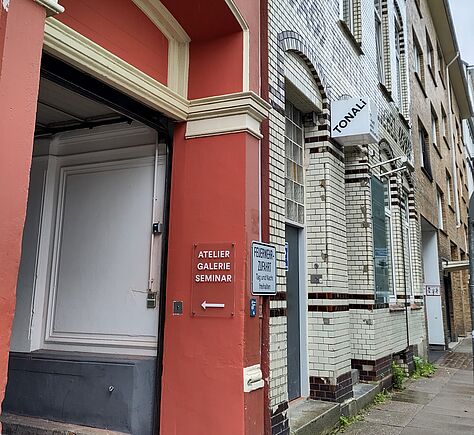 Ein roter Torbogen mit weißen Absätzen. An der Wand ein Schild mit der Aufschrift ATELIER GALERIE SEMINAR in weißen Buchstaben. Das Nachbarhaus hat eine weiße Fassade mit dunkelbraunen Ziegelsteinverzierungen