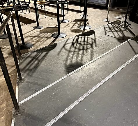 Im Vordergrund verlaufen zwei schmale Podeste mit silberfarbenen Kanten über eine leicht erhöhte Fläche. Links ist ein Handlauf aus Metall montiert. Im Hintergrund stehen mehrere hohe, runde Stehtische auf einem helleren Holzboden. 