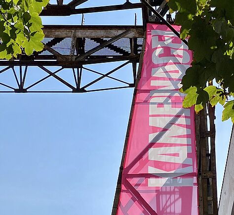 In den blauen Himmel fotografiert. Ein Teil des pinkfarbenen Banners mit der Aufschrift Kampnagel. Oben im Bild ein Ausschnitt einer alten Krankonstruktion