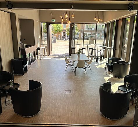 Heller, moderner Raum mit Holzboden. In der Mitte steht ein rechteckiger Tisch mit mehreren Stühlen. An den Seiten und in den Ecken stehen schwarze, runde Sessel, jeweils an kleinen Beistelltischen. Die Wand auf der rechten Seite ist fast komplett aus Glas. Im Hintergrund gibt es hohe Fenster und eine große, geöffnete Doppeltür mit Blick nach draußen auf einen Platz mit Bäumen und Gebäuden. An der linken Wand steht eine Theke mit Barhockern. An der Decke mehrere Glühbirnenlampen.