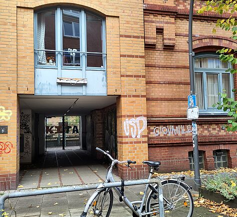 Die Hausfassade aus rötlich-gelben Backsteinen mit zwei großen Fenstern. Zentral die Hofeinfahrt, links und rechts Graffiti-Gemälde an den Wänden, am Ende ein Tor, darauf steht in großen Buchstaben "KITA". Im Vordergrund steht ein Fahrrad, das an einem Metallgeländer angeschlossen ist. Rechts daneben steht eine Straßenlaterne mit einem blauen Schild für Parkplätze und einem darunter angebrachten weißen Zusatzschild, das auf einen Rollstuhlplatz hinweist. Am rechten Rand ragt ein Baum mit grünem Laub ins Bild. Auf dem Boden und am Pflanzenbeet rechts liegen verstreut gelbe und braune Herbstblätter.