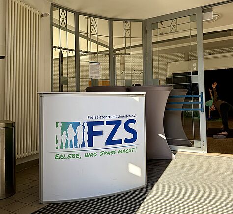 Vorn steht ein Tresen mit dem Logo „FZS“. Dahinter sind große Glasfenster und eine Glastür zu sehen. Durch die Tür sieht man einen Raum, in dem eine Person Sport oder Yoga macht.