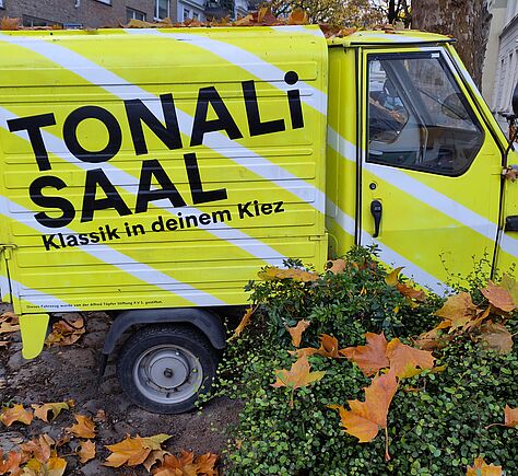 Gelb gestrichenes, kleines dreirädriges Fahrzeug (eine Ape) steht auf einem Platz, der mit Herbstlaub bedeckt ist. Darauf steht in schwarzen, großen Buchstaben: "TONALi SAAL". Darunter in kleinerer Schrift: "Klassik in deinem Kiez". Das Fahrzeug trägt schräg verlaufende weiße Streifen.