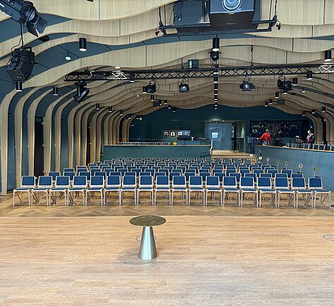 Der Saal mit vielen Reihen blauer Stühle und einem Holzfußboden. Das Foto ist von der Bühne aus in den Saal hinein aufgenommen. Wände und Decke haben eine wellenförmige Struktur aus Holz. An der Decke hängen mehrere große, schwarze Scheinwerfer. In Blickrichtung sind rechts an der Seite Plätze zu sehen, die von der oberen Ebene aus erreicht werden.