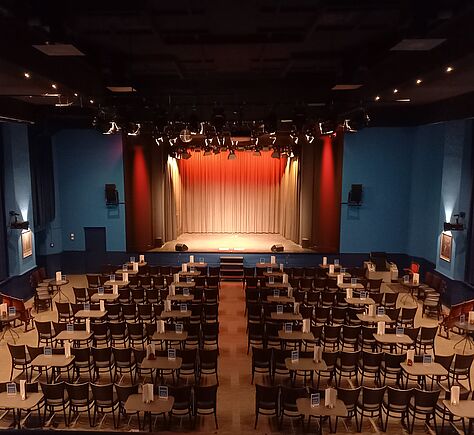 Blick auf die Bühne, hinten ein gelb-roter Vorhang. Der Saal ist in Reihen bestuhlt, dazwischen stehen kleine Tische.