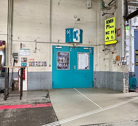 Türkisfarbene Doppeltür mit der großen weißen Ziffer '3' über dem Eingang in einer Industriehalle. Auf beiden Tür sind Plakate, ebenso links und rechts an den hellen Wänden. Über der Tür befindet sich ein türkisfarbenes Schild mit weißem Symbol. Rechts hängt ein gelbes Schild mit den Aufschriften 'KMH', 'P1', 'WC' sowie Piktogrammen für Wickelraum und barrierefreie Toilette. Der Boden ist grau und beige, mit einer weißen Bodenleitlinie für Blinde, die auf die Tür zuführt und mit drei Streifen auch weiter Richtung KMP, P1 und WC. Die Wände sind weiß und grau gestrichen, links befindet sich ein Stehtisch.