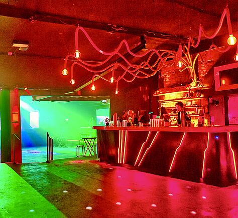 Eine Bar mit leuchtend roten LED-Streifen und einer auffälligen Deckeninstallation aus Kabeln und Glühbirnen. Dahinter befindet sich ein offener Raum mit grüner und blauer Lichtinstallation. Der Boden ist aus Beton.