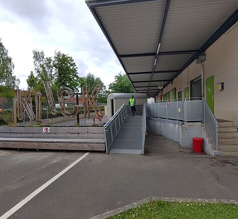 Am Gebäude ist rechts eine Steintreppe mit sechs Stufen und einem Handlauf auf der linken Seite. Mit etwas Abstand ist links neben der Treppe eine Rampe aus Metall mit Handläufen rechts und links. Von Treppe und Rampe geht es rechts zur Eingangstür.