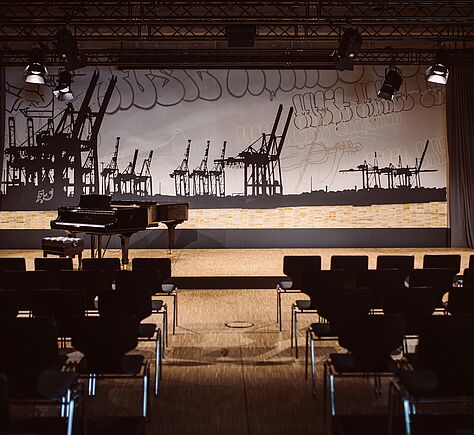 Eine Bühne in einem Saal mit einem schwarzen Flügel und Sitzreihen im Vordergrund. Vor der Bühne stehen viele dunkle Stühle in Reihen, getrennt durch einen Mittelgang; der Boden helles Holz.  Die Rückwand der Bühne ist mit einer großen, grafischen Silhouette von Hafenkränen gestaltet, schwarz vor einem hellen Hintergrund. Darüber sind großflächige, geschwungene Linien und skizzenhafte Formen in dunkler Farbe zu sehen. Oben hängen mehrere schwarze Bühnenscheinwerfer und eine Traverse; rechts ist ein dunkler Vorhang. 