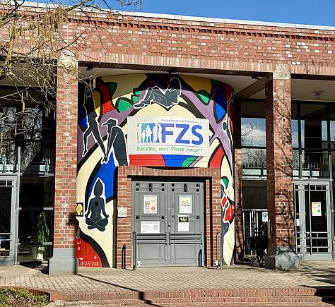 Ein Gebäude aus rotem Backstein mit einem Eingang in der Mitte. Über der Tür hängt ein Schild mit der Aufschrift „FZS“. Rund um den Eingang ist eine große, bunte Wandmalerei mit verschiedenen Figuren zu sehen.