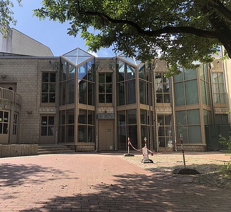 Das Gebäude hat eine Fassade aus Glas und hellem Stein. In der Mitte ist eine Eingangstür aus Glas. Vor dem Gebäude ein gepflasterter Platz mit großen Bäumen. Das Foto ist an einem sonnigen Sommertag aufgenommen worden.