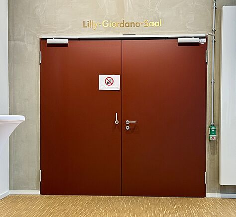 Eine große doppelte Tür aus rotem Metall mit glatter Oberfläche und silbernen Griffen. Über der Tür steht in goldenen Buchstaben „Lilly-Giordano-Saal“. Die helle Betonwand und der Holzboden wirken ruhig und schlicht, der Raum wirkt modern und ordentlich.