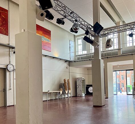 Glänzender Linoleum-Fußboden, im Hintergrund ein großes bodentiefes Fenster. Hinten in der Ecke ein Kühlschrank. An der linken Wand hängen im obersten Drittel 2 große abstrakte Bilder in Rot-Tönen. Auf der linken Seite 2 eckige Säulen, an der Decke hängt Gestänge mit daran befestigten Scheinwerfern und Boxen.