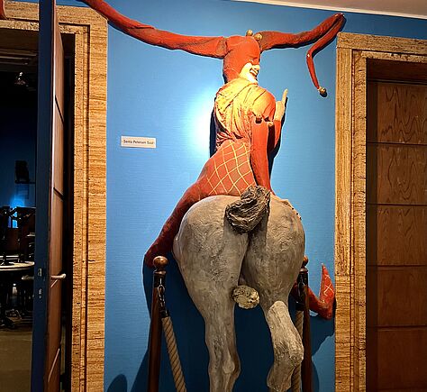 Auf der blauen Wand ist Skulptur angebracht zu sehen, ist das Hinterteil eines Pferdes, auf die ein Harlekin sitzt, der den Stinkefinger zeigt. Links und rechts von dieser Raum hohen Skulptur sind im Anschnitt, zwei Türen zu sehen.