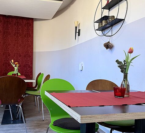 Ein kleiner Café- oder Bistro-Sitzbereich mit Tischen und Stühlen in Rot-, Grün- und Holztönen. Im Vordergrund steht ein Tisch mit einem roten Tischläufer; darauf steht eine Glasvase mit einer einzelnen rosa Tulpe und ein rotes Glas mit Kerze. Im Himntergrund ein weiterer, genauso ausgestatteter Tisch. Rund um die Tische stehen Stühle mit grünen und braunen Lehnen. Die Rückwand des Raumes ist dunkelrote. Rechts an der Wand hängt eine Wandleuchte und ein rundes Wandregal mit Deko-Objekten.