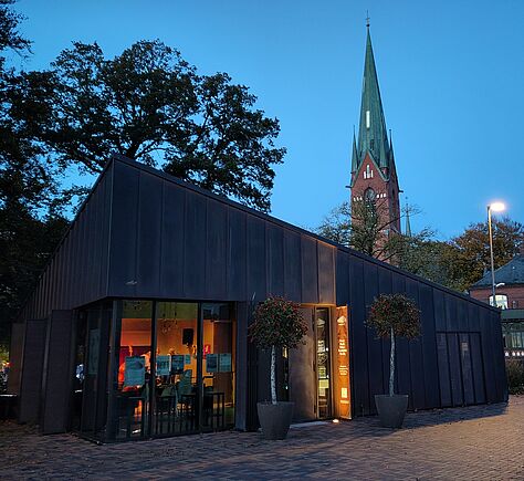 Modernes, dunkles, einstöckiges Gebäude. Durch die großen Fenster sind innen Personen zu erkennen. Vor dem Eingang stehen zwei Bäume in großen Töpfen. Im Hintergrund ragt der hohe, spitze Turm einer Backsteinkirche in den Abendhimmel. Eine Straßenlaterne beleuchtet den Platz.