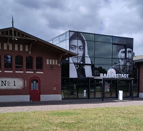 Altes Backstein-Gebäude mit modernem Glas-Erweiterungsbau. Links steht ein einstöckiges Backsteingebäude mit schrägem Dach. Die Fassade hat mehrere hohe Rundbogenfenster. An der Wand ist ein hell abgesetztes großes Schild mit der Aufschrift „Nº 1“. Rechts schließt ein würfelförmiger Glasbau an, dessen Front vollständig aus Glasflächen besteht. Auf dem Glas sind zwei große schwarz‑weiße Porträtfotos angebracht. Über dem Eingang ist ein rundes Logo mit einer stilisierten, bunten Kugel und darunter das Wort „BALLINSTADT“ in weißen Großbuchstaben. Im Vordergrund eine größere Rasenfläche.