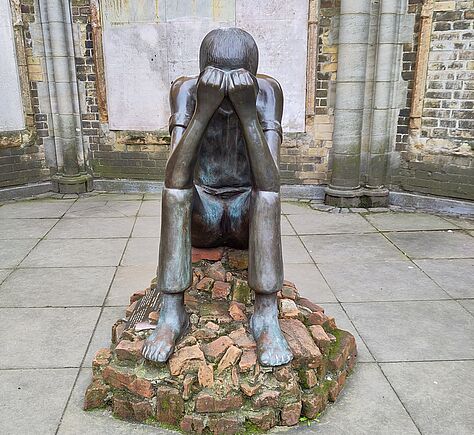 Eine sitzende Figur aus dunklem Metall mit glatter, leicht glänzender Oberfläche. Die Person hält den Kopf in den Händen, die Beine sind angewinkelt und die Füße stehen auf einem Sockel aus unregelmäßigen Ziegelsteinen. Die Skulptur steht in einem Innenhof mit Steinboden und alten Mauern.